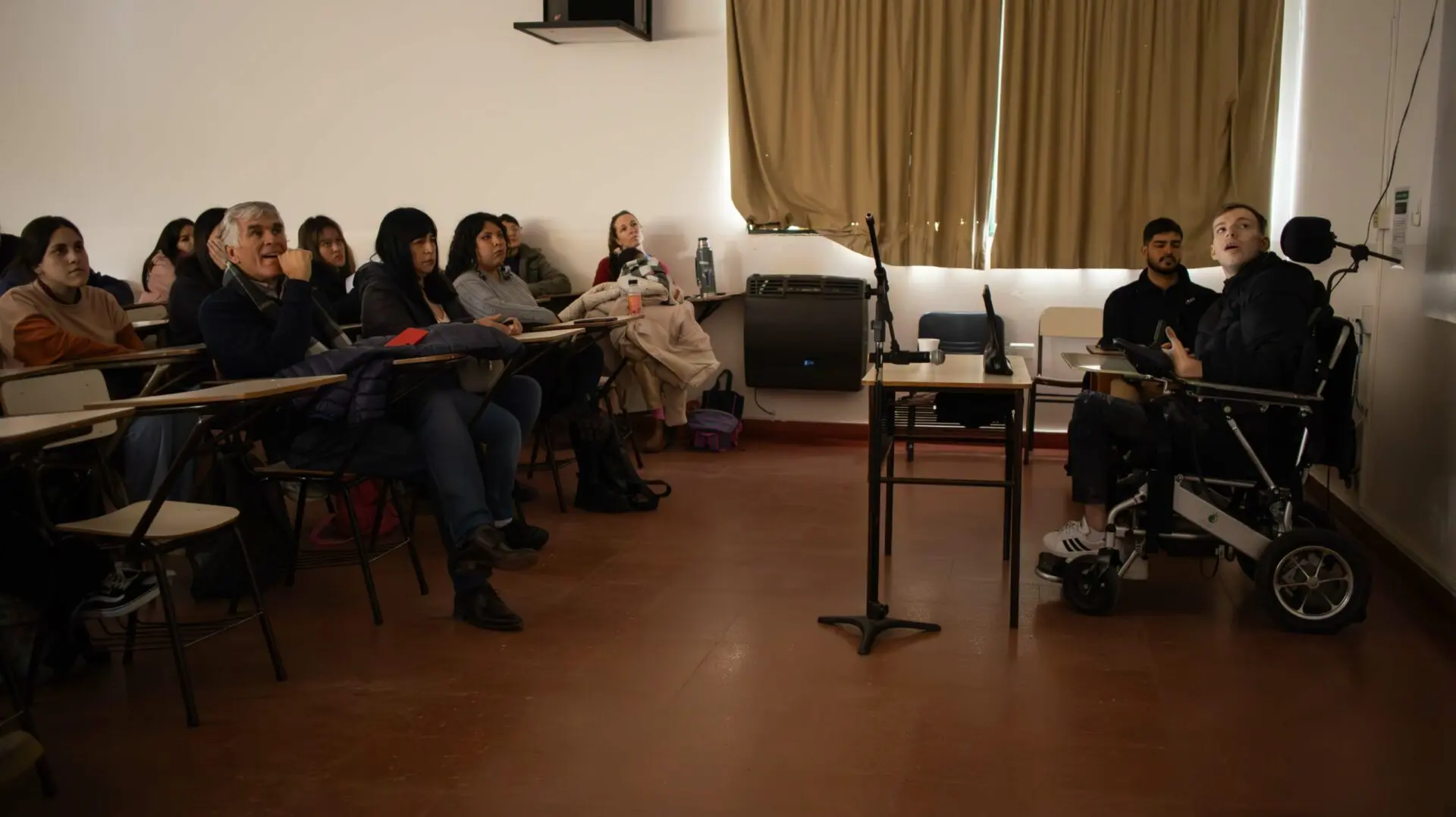 Iván Davidovich dando una charla sobre comunicación accesible y CAA en un aula, acompañado por su dispositivo en silla de ruedas, frente a estudiantes atentos.