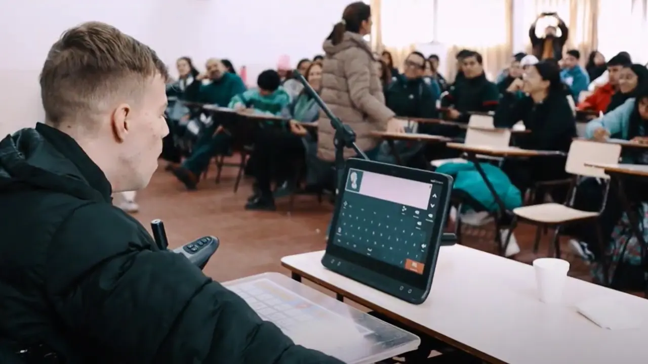 Iván Davidovich dando una charla en un aula inclusiva, utilizando un dispositivo de comunicación aumentativa frente a estudiantes.