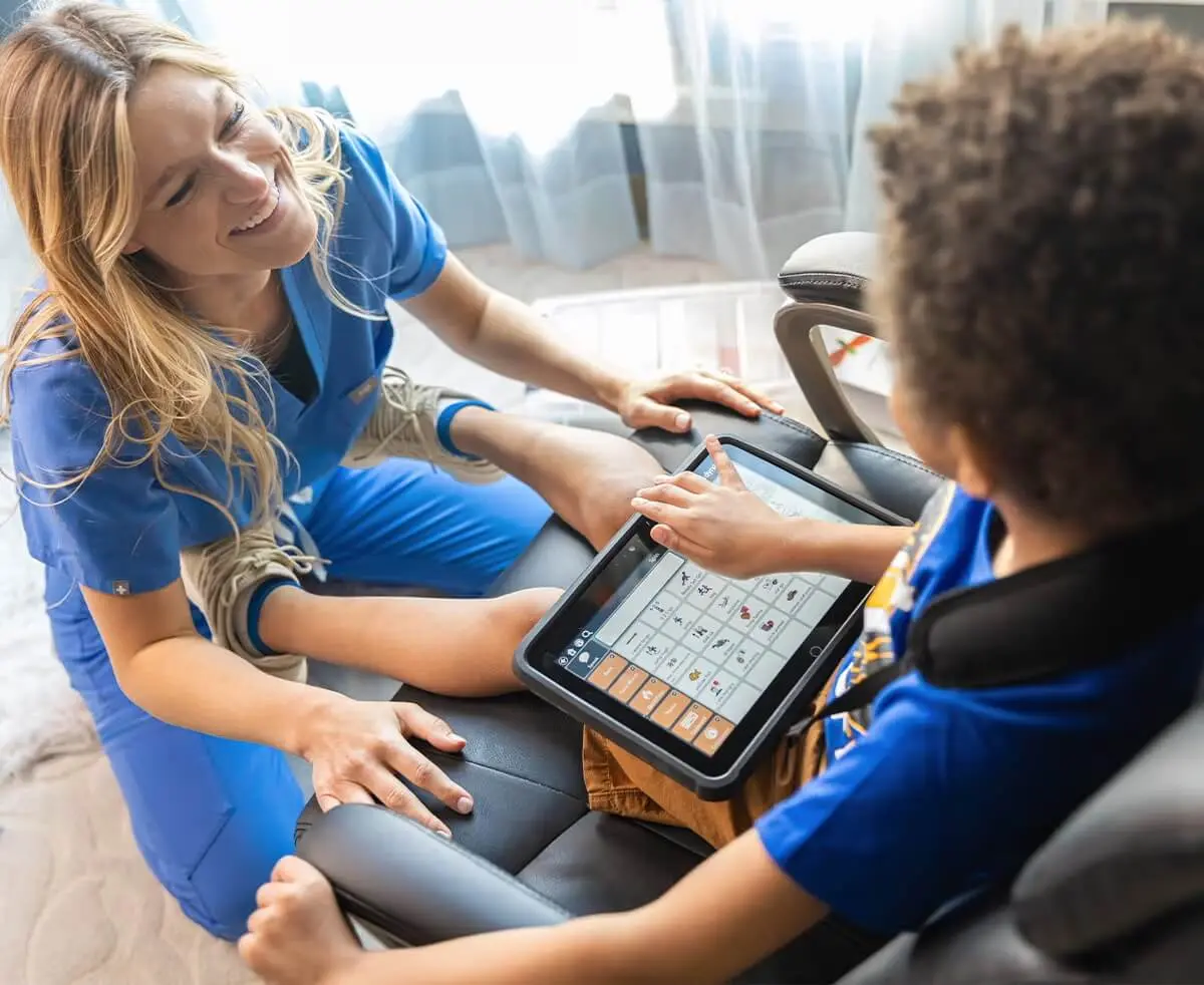 Niño usando un dispositivo de comunicación aumentativa en una tablet mientras una terapeuta lo acompaña sonriente.