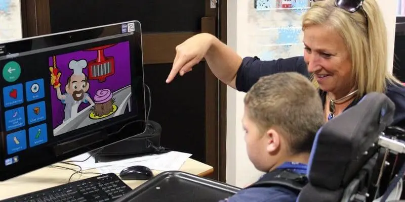 Mujer acompaña a un niño con discapacidad a usar un software de comunicación aumentativa en la computadora.