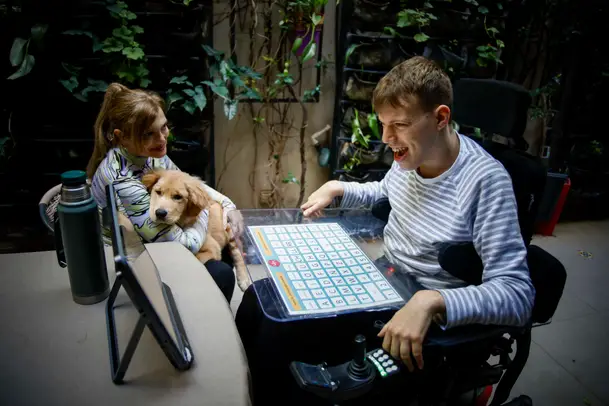 Iván Davidovich utilizando un tablero de Comunicación Aumentativa y Alternativa junto a su mamá y un perro, en una escena de apoyo y autonomía.